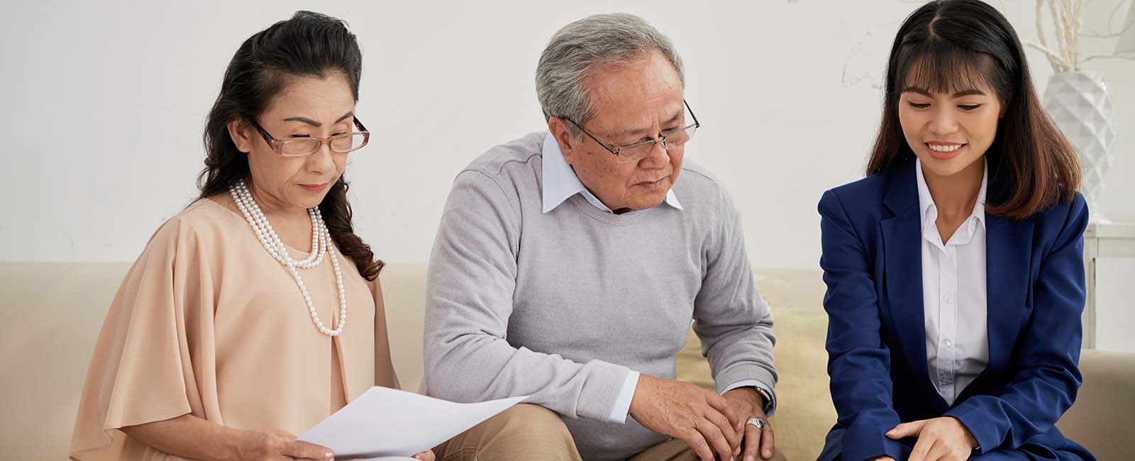 A woman in glasses holds papers while sitting on a sofa next to an older man in a gray sweater and another woman in a blue blazer. They are focused on the documents, suggesting a discussion or consultation in a comfortable setting with a white wall in the background.