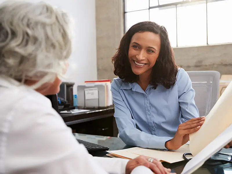 A woman in a light blue blouse is seated at a desk, smiling and showing a folder to an older woman with gray hair. They are in an office setting with files and a large window in the background, suggesting a professional or collaborative meeting.