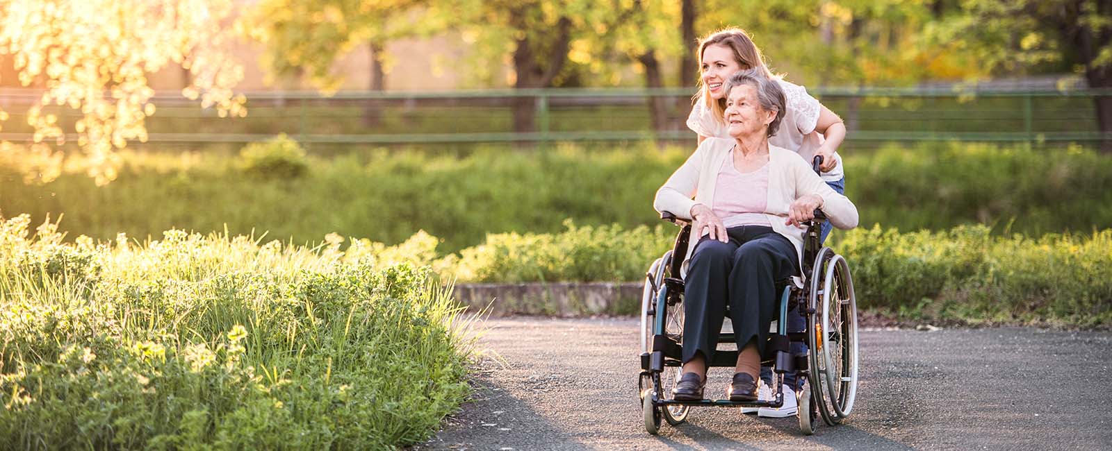 A young woman happily pushes an elderly woman in a wheelchair along a sunlit pathway. They are surrounded by lush greenery and trees, with sunlight filtering through the leaves. Both appear content and are enjoying the peaceful, outdoor setting.