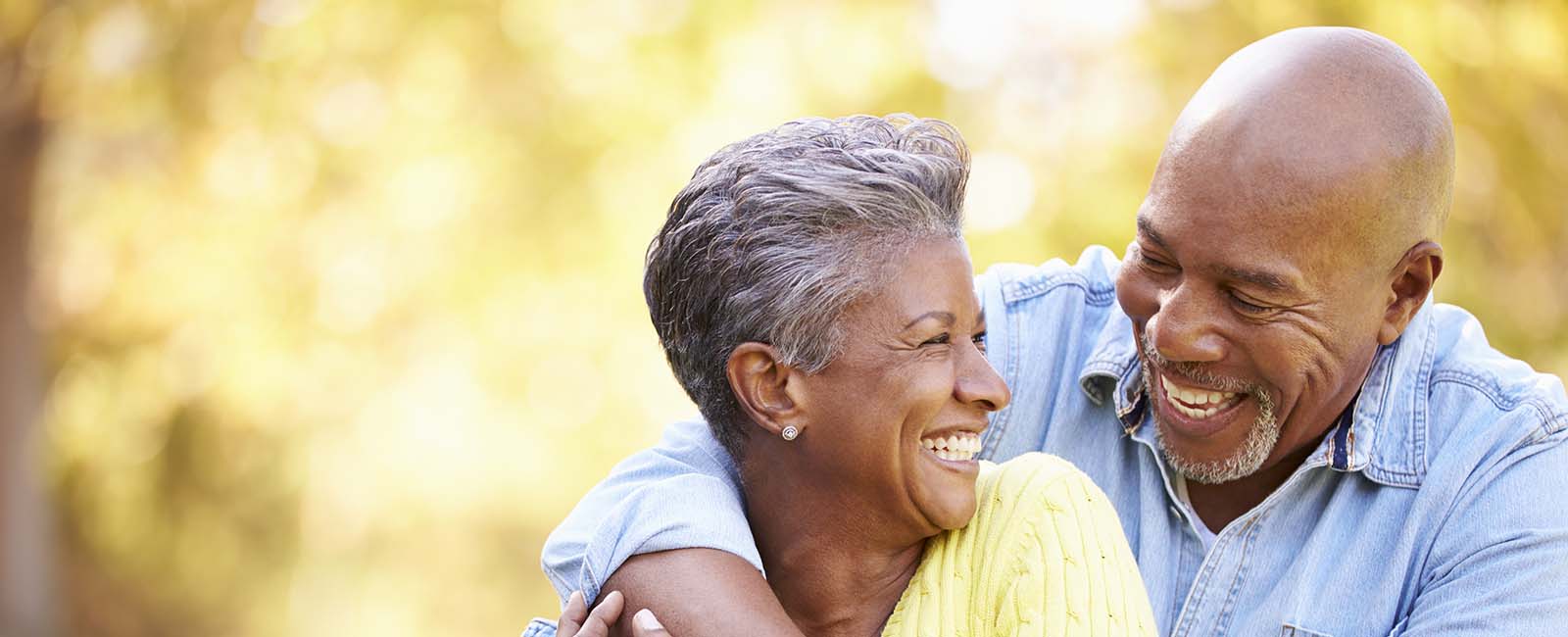 An older couple stands outdoors, warmly embracing and smiling at each other. The woman wears a yellow shirt, and the man is in a denim shirt. The background features a softly blurred, sunlit natural setting, creating a cheerful and tender atmosphere.
