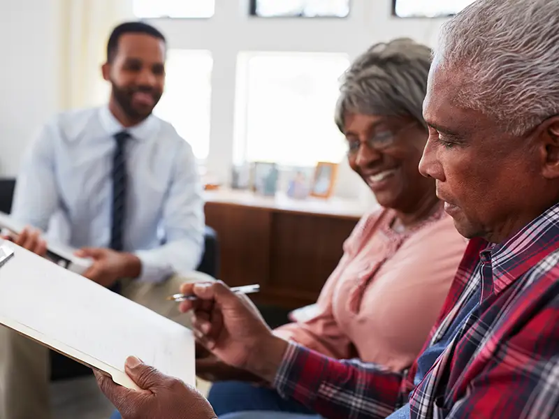 An elderly couple sits together, reviewing documents on a clipboard. The man wears a plaid shirt, while the woman in a pink blouse smiles and holds a pen. A man in a white shirt and tie, blurred in the background, appears to be explaining something. Sunlight streams in through a window.