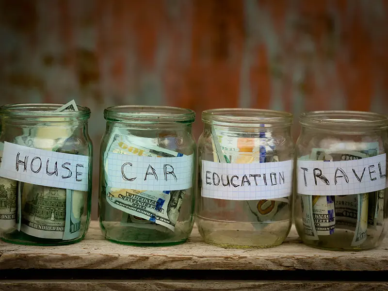 Four glass jars labeled “HOUSE,” “CAR,” “EDUCATION,” and “TRAVEL” filled with various U.S. dollar bills sit on a wooden surface, symbolizing life planning and thoughtful saving, with a blurred brown and orange background.