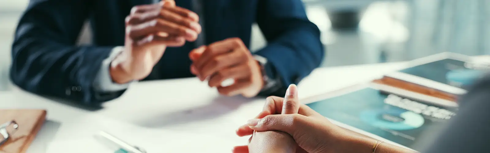 Two people sit across a table in a professional setting, discussing audit preparation. One gestures while the other listens, hands clasped. Documents and charts on the white table suggest they're reviewing audit steps in this focused business meeting. Faces are not visible; focus is on hands.