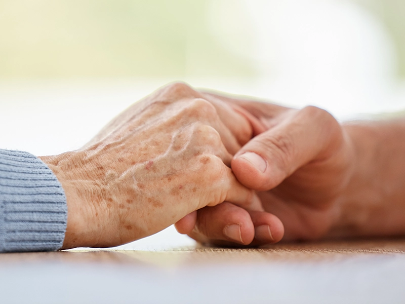 A close-up of two hands clasped together on a table—one older, one younger. The scene subtly reflects the emotional moments that can arise when you probate an estate in Virginia. The background is softly blurred, with the older person in a light blue sweater.