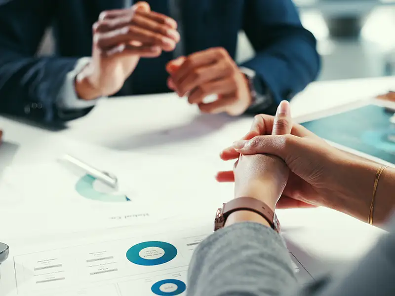 Two people in business attire sit at a desk reviewing accounting charts and graphs. One gestures while the other’s hands are clasped. A pen and a tablet with a blue-toned chart highlight their focus on financial management documents.