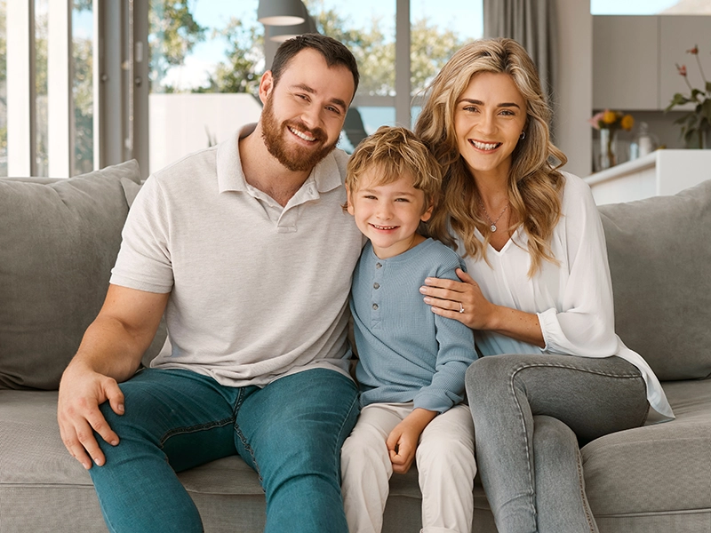 A smiling man, woman, and young boy sit together on a gray couch in a bright living room. As they relax, they discuss the definition of a Will, ensuring their family's future is protected in a warm, sunlit space.