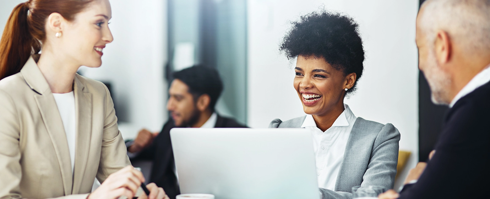 Three professionals sit at a table in an office, engaged in conversation. A woman in a gray suit smiles broadly at a woman in a beige suit. A man sits beside them. A laptop is open in front. Another person works in the blurred background.