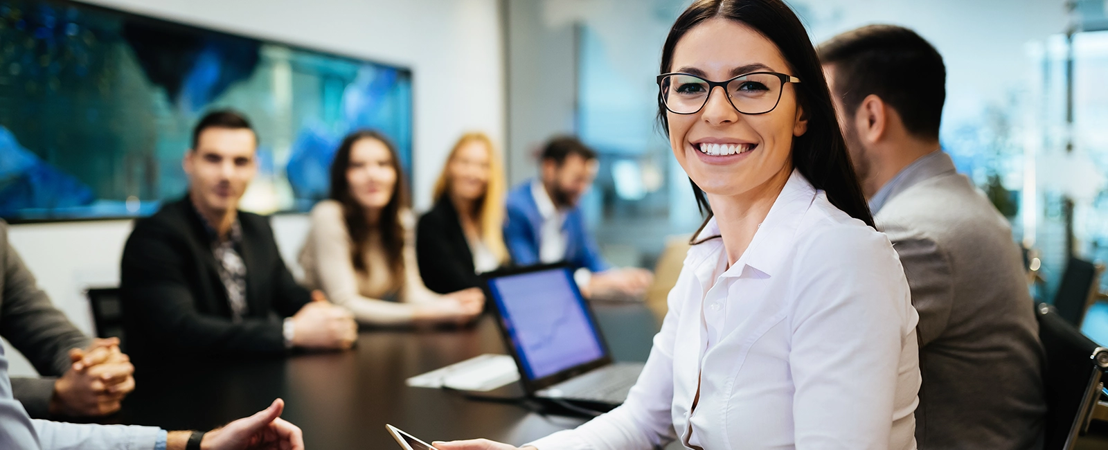A woman in glasses and a white shirt smiles at the camera while sitting at a conference table with colleagues. Several people are blurred in the background, seated around the table with a laptop and documents visible. The scene is bright and professional.