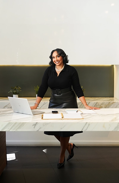 A woman in a black dress and heels stands smiling behind a marble desk with an open laptop, papers, and pens. She wears a pearl necklace and bracelet. The modern office has plants and a sleek, minimalist design with soft lighting.